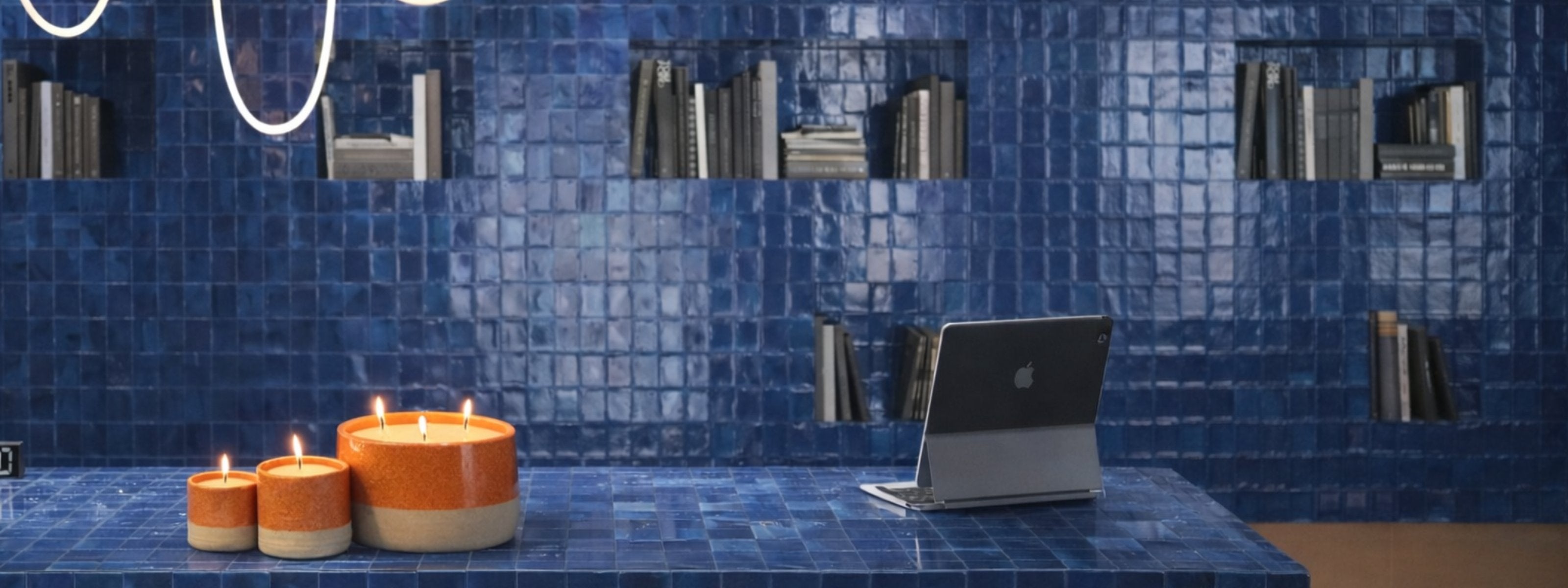 Bougies parfumées Maison Tassan en céramique posées sur une table en carrelage bleu dans un intérieur moderne avec bibliothèque murale et éclairage design.